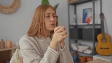 Oturma odasında elleri sımsıkı bağlı, gözleri kapalı, rahat bir süveter giymiş, etrafı kitaplar ve gitarla çevrili, huzurlu bir evde dua eden bir kadın..