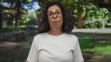 Orta yaşlı İspanyol bir kadın yeşil bir açık hava parkında gülümsüyor, beyaz bir gömlek giyiyor, güneş ışığı ve ağaçlarla çevrili, canlı ve neşeli bir atmosfer yaratıyor..