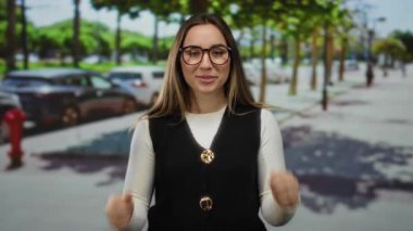 Güneşli bir sokakta genç bir kadın, kendine güvenen bir ifade ve günlük kıyafetle gözlük takıyor, park edilmiş arabalar ve şehir manzarasında yapraklı yeşil ağaçlarla çevrili..