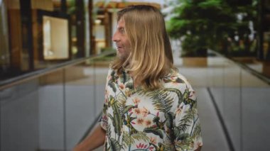 Sarışın ve uzun saçlı bir adam gün ışığında bina koridorunda cam parmaklıklara işaret ediyor..