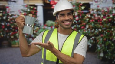 Kask ve güvenlik yeleği giyen adam kahve demliğini elinde tutuyor ve binanın önünde gülümsüyor; sabah rutin mutluluğu.