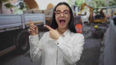 Kırmızı gözlüklü beyaz laboratuar önlüklü İspanyol kadın inşaat alanında duruyor ve devam eden faaliyetlere şaşırmış bir şekilde bakıyor..