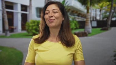 Sarı bluz giyen İspanyol orta yaşlı kadın, şehir sokağında palmiye ağaçları ve bina cephesiyle dolu sıcak bir gülümsemeyle çifte onay veriyor; onay pozitifliği iyimserliği teşvik ediyor.
