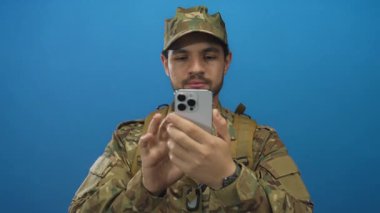 Kamuflaj üniformalı genç İspanyol adam mavi stüdyoda parmak uçlarıyla akıllı telefon ekranını dinliyor;.
