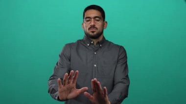 Gri tişörtlü, yeşil arka planda elleri havada, gözlüklü genç bir adam, bir uyarı ya da tereddüt işareti yapıyor..