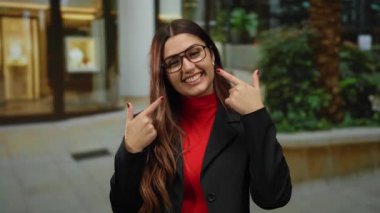 Kalabalık bir sokakta gülümseyen ve gülümseyen bir kadın, açık hava ortamına güvendiğini gösteren, siyah takım elbiseli ve gözlüklü kırmızı bluzlu bir kadın..