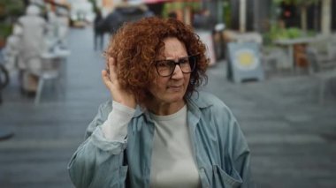 Orta yaşlı, kızıl saçlı ve gözlüklü bir kadın kalabalık bir şehir caddesinde merakla ve nişanlanmayla ilgili bir jest yapıyor..