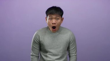 Şaşırmış bir ifadeye sahip genç bir adam, izole bir mor arka plana karşı duruyor, gri bir gömlek giyiyor, bir anlık şok ve duyguyu özetliyor..