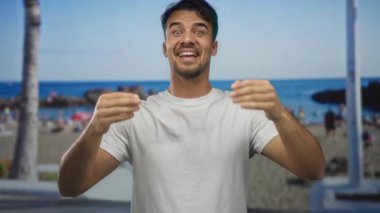 Palmiye ağaçları ve canlı bir açık hava sahnesi ile deniz kıyısında gülümseyen genç İspanyol adam, arka planda parlak mavi bir deniz ile neşeli bir yaz anını yakalıyor..