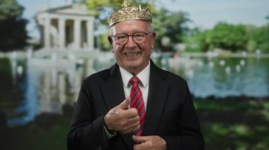 Parkta taç giyen kıdemli bir adam, elleri ve elleri ile bir yandan da takım elbise giymiş, manzaralı bir arka plana karşı hazırlanmış neşeli bir davranışı vurguluyor..