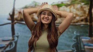 Güneşli bir plajda şapkayla poz veren, deniz kenarında yaz tatili sırasında gençlik neşesi sergileyen bir kadın..
