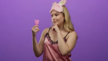 Pembe gecelikli kadın elinde mor arka planda tuhaf bir ifade olan adet bardağı tutuyor.