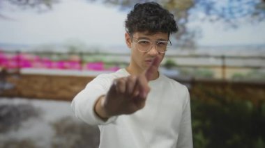 Beyaz süveterli ve yuvarlak gözlüklü genç İspanyol adam yoğun caddede kamerayı işaret ediyor; şüphecilik.