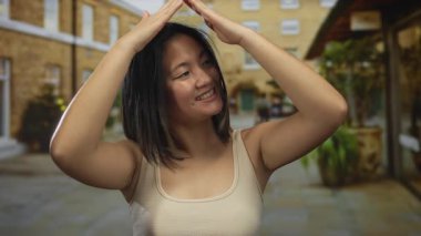 Açık havada gülümseyen bir kadın güneşli bir günde şehir merkezinde tuğla binalarla çevrili bir caddede çatı jesti yapıyor..