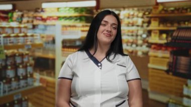 Kendine güveni tam genç bir kadın süpermarkette gülümsüyor. Etrafı ürün raflarıyla çevrili. Perakende satış ortamında kıvrımlı bir figür sergiliyor..