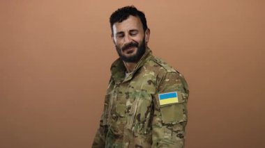 Ukrayna bayraklı askeri üniformalı İspanyol bir adam, turuncu bir arka plana gülümsüyor..