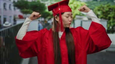 Kırmızı mezuniyet elbisesi içindeki kendine güvenen kadın, başarı ve başarıyı simgeleyen şehir sokaklarında muzaffer bir poz veriyor..