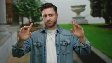 Sakallı genç adam şehir dışında bir sokak ortamında elini kaldırıyor, kot ceket giyiyor, düşünceli bir bakışla jest yapıyor, yemyeşil ve şehir mimarisi ile çevrili..