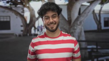 Sakallı ve gözlüklü genç adam arka planda ağaçlar ve binalar olan bir sokakta gülümsüyor kırmızı çizgili bir gömlek giyiyor..