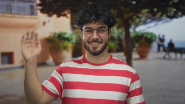 Sırıtan sakallı genç bir adam çizgili bir tişörtle sokakta ağaçlarla ve arka planda bir bankla durup sıradan ve dostane bir atmosfer yaratıyor.