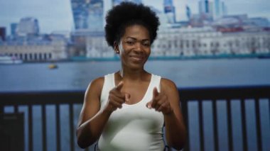 Arka planda şehir silüeti olan kamerayı gösteren bir kadın, açık şehir gezinti alanı, beyaz bluz giyen, neşeli bir ifade sergileyen, canlı bir şehir atmosferi tasvir eden bir kadın..