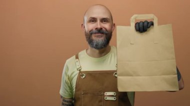 Kahverengi arka planda sakallı bir adam, önlük ve siyah eldiven giyiyor, yiyecek dağıtım hizmeti sunuyor, çevreye duyarlı ambalajları işaretliyor..