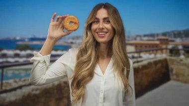 Sarışın kadın, deniz kenarındaki bir balkonda gözünün yanında donut tutarken gülümsüyor..