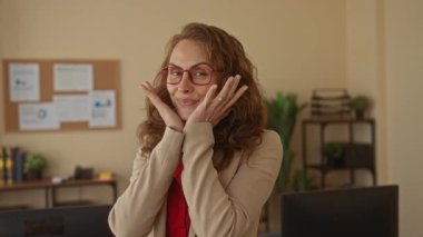 Ofis ortamında güvenle gülümseyen, gözlüklü ve ceketli, etrafı kitaplarla ve bitkilerle çevrili, çalışma ortamında mutluluk ve profesyonellik taşıyan bir kadın..