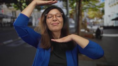 Mavi ceketli kadın, elleri ağaçlarla dolu bir caddede yüzünü çerçeveliyor..