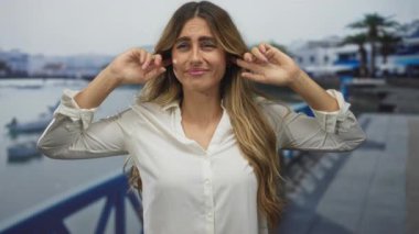 Beyaz bluzlu genç sarışın kadın, güneşli bir yat limanında, demirli teknelere tepeden bakan kulaklarını kapatıyor..