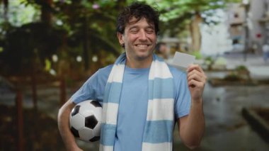 Şehir dışında elinde futbol topu ve kart tutan İspanyol bir adam şehir merkezinde fular takıyor..