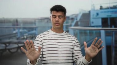 Bir yolcu gemisinin güvertesinde, elleri havada, çizgili gömlek giymiş, arka planda bulanık oturma ve gemi korkulukları olan bir adam..