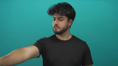 Sakallı genç adam mavi arka plana karşı küçümseyici bir jest yapıyor. Ciddi görünüyor ve odaklanmış. Hareketi, karar ya da reddedilmeyi ima ediyor.