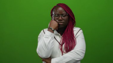 Yorgun yüzlü bir kadın yeşil arka plana karşı duruyor, gözlük takıyor ve beyaz bir kazak giyiyor, örgülü saç stilini ve derin düşüncelere dalmış ruh halini gösteriyor..