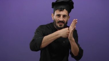 Siyah üniformalı İspanyol erkek şef, mor bir arka plana karşı el çırpıyor, neşeli ve enerjik bir ifade sergiliyor..