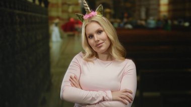 Sarışın kadın tek boynuzlu at diademi takmış Katolik kilisesinde gülümsüyor kolları çapraz ve neşeli bir ifadeyle.