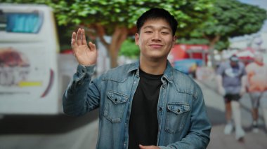 Kot ceketli genç Çinli adam ağaçlar ve yayalar kameraya el sallıyor..