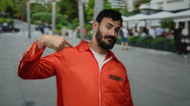 Hapishane tulumu giyen Hispanik bir adam işlek bir caddede, kentin dış manzarasıyla çevrili bir sırıtışla kendine bakıyor..