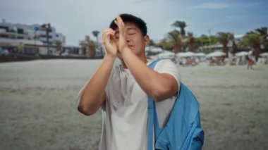 Genç Çinli adam deniz kenarında esniyor, beyaz gömlek ve mavi sırt çantası giyiyor, açık deniz manzarasının keyfini çıkarıyor, öğrenci rahatlığını ve huzurlu deniz manzarasını yakalıyor..