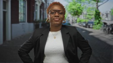 Genç Afro-Amerikalı kadın, binalar ve park edilmiş arabalarla çerçevelenmiş kentsel sokak sahnesinde elleri kalçalarının üzerinde duruyor..