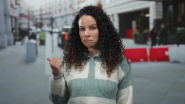 İşlek bir caddede baş parmağıyla işaret veren kadın bir an düşünüp taşınıyor ve şehir dışında bir arka planda yüzünü gösteriyor..