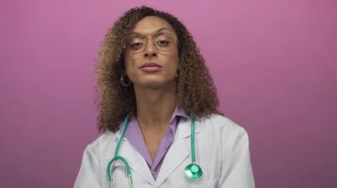 Yeşil steteskoplu beyaz önlüklü kadın doktor pembe stüdyo duvarına karşı iki parmağını da kaldırıyor..