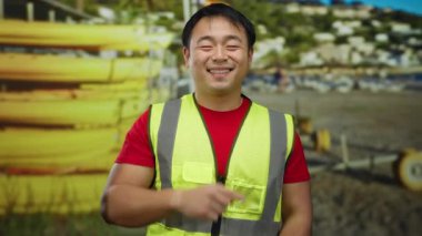 Güvenlik yeleği giyen Asyalı bir adam, arka planda canlı sarı ekipmanlar bulunan bir deniz kenarı şantiyesinde güvenle gülümsüyor..