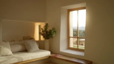 Güneşli pencereli, yumuşak yastıklı ve yeşillikli lüks bir Akdeniz evinin içi huzurlu ve rahat bir yaşam alanını vurguluyor..