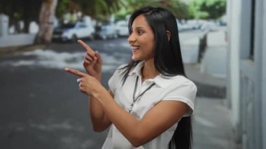 Güneşli bir sokakta güvenle gülümseyen genç bir kadın park etmiş arabaların ve yeşil ağaçların arka planında, elleriyle işaret eden, beyaz gömlek ve isim kartı takan.