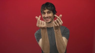 Rahat duruşu olan genç adam kırmızı stüdyoda parmak kalp hareketi yapıyor, kameraya sıcak bir şekilde gülümsüyor;.