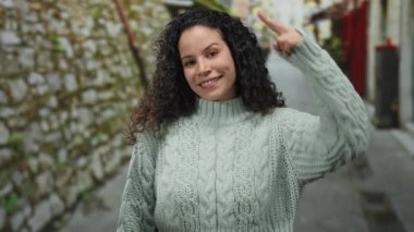 Sokakta gülümseyen, süveter giyen, elleri kafasında şakacı hareketler yapan, kıvırcık saçlı, taş duvarında yeşillik olan canlı şehir ortamında İspanyol kökenli bir kadın..