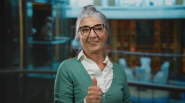 Otelin içindeki gri saçlı kadın gülümseyip işaret ederken, gözlük takarken, yeşil bir kazak giyerken, coşku ve pozitifliği iletirken başparmak kaldırıyor..