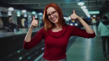 Kapalı bir tren istasyonunda kırmızı tişört ve gözlük takan, neşeli bir şekilde gülümseyen bir kadın.