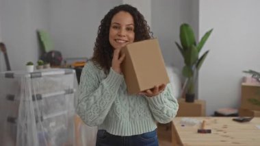 Yeni evinde elinde karton bir kutuyla gezen genç bir kadın. Oturma odasında içi boş eşyalar ve bitkilerle birlikte. İçeriden heyecan ve taze bir başlangıç hissi yayıyor..